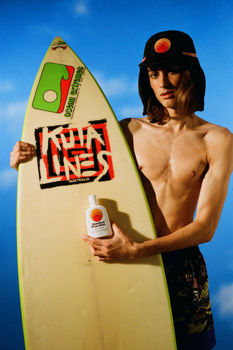 Sun block SPF 50 bottle held by a surfer with a board and wearing a sun-protective hat under a clear blue sky.
