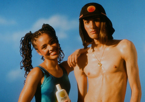 Australian made sunscreen held by a smiling woman next to a man wearing a sun-protective hat under a clear blue sky.