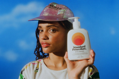 Broad spectrum sunscreen SPF 50 bottle held by a model wearing a pink iridescent cap against a bright blue sky backdrop.