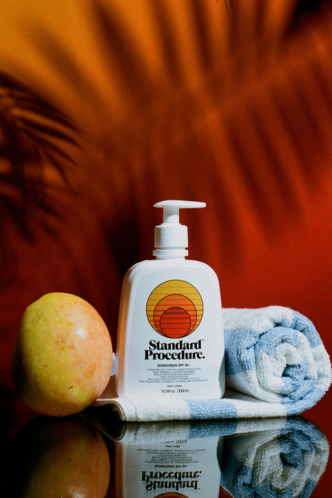 Sunblock SPF 50 pump bottle beside a fresh mango and rolled beach towel on a reflective surface.
