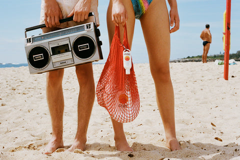 Sun cream SPF 50 clipped to a red mesh beach bag held by a woman standing barefoot on the sand.