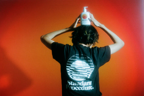 Sunblock cream SPF 50 bottle balanced on a person's head wearing a black Standard Procedure shirt against an orange gradient background.