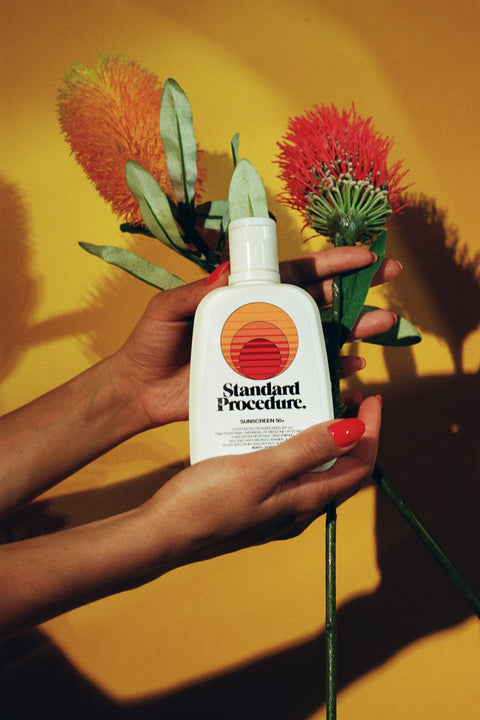Vitamin C with sunscreen bottle held by hands alongside red and orange flowers against yellow backdrop.