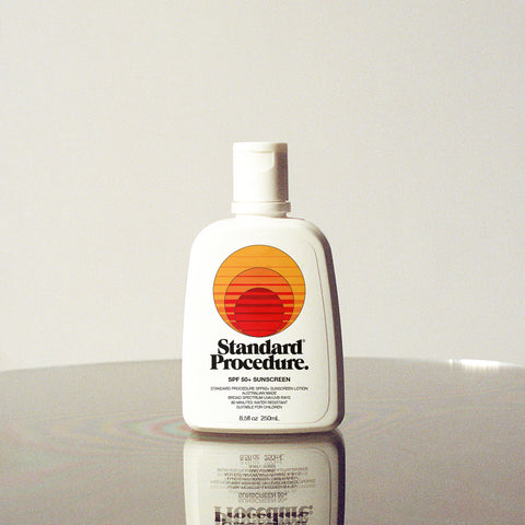 SPF 50 sunscreen bottle with pump top and Standard Procedure logo standing on a reflective white surface.
