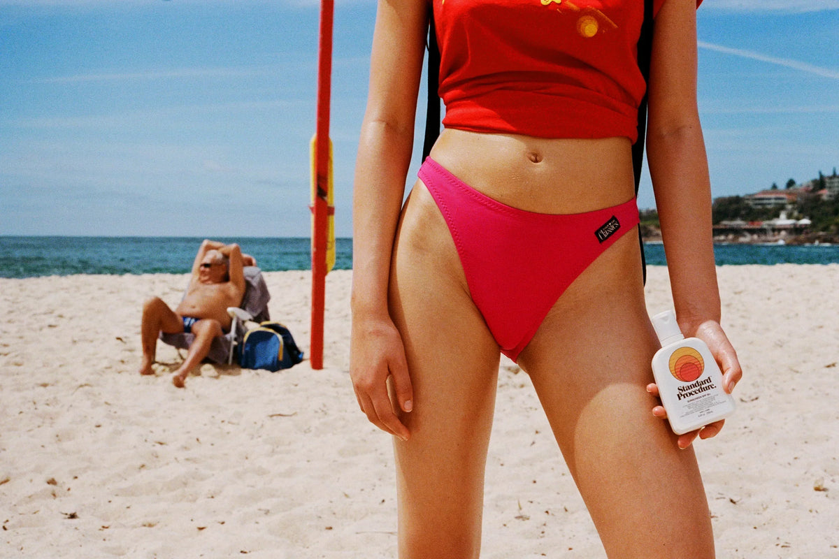 Person on a sunny beach holding a bottle of Standard Procedure SPF 50 sunscreen.