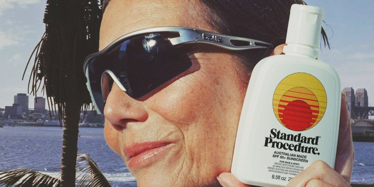 Person in sports sunglasses holding a Standard Procedure SPF 50+ sunscreen bottle outdoors.