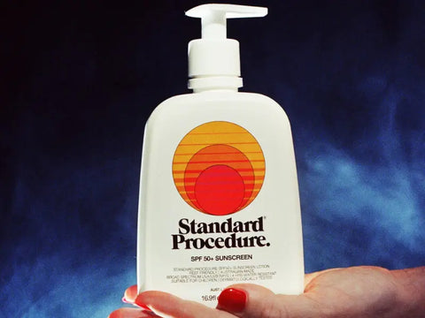 SPF sunscreen bottle of Standard Procedure SPF 50+ held in a hand against a dark blue background.