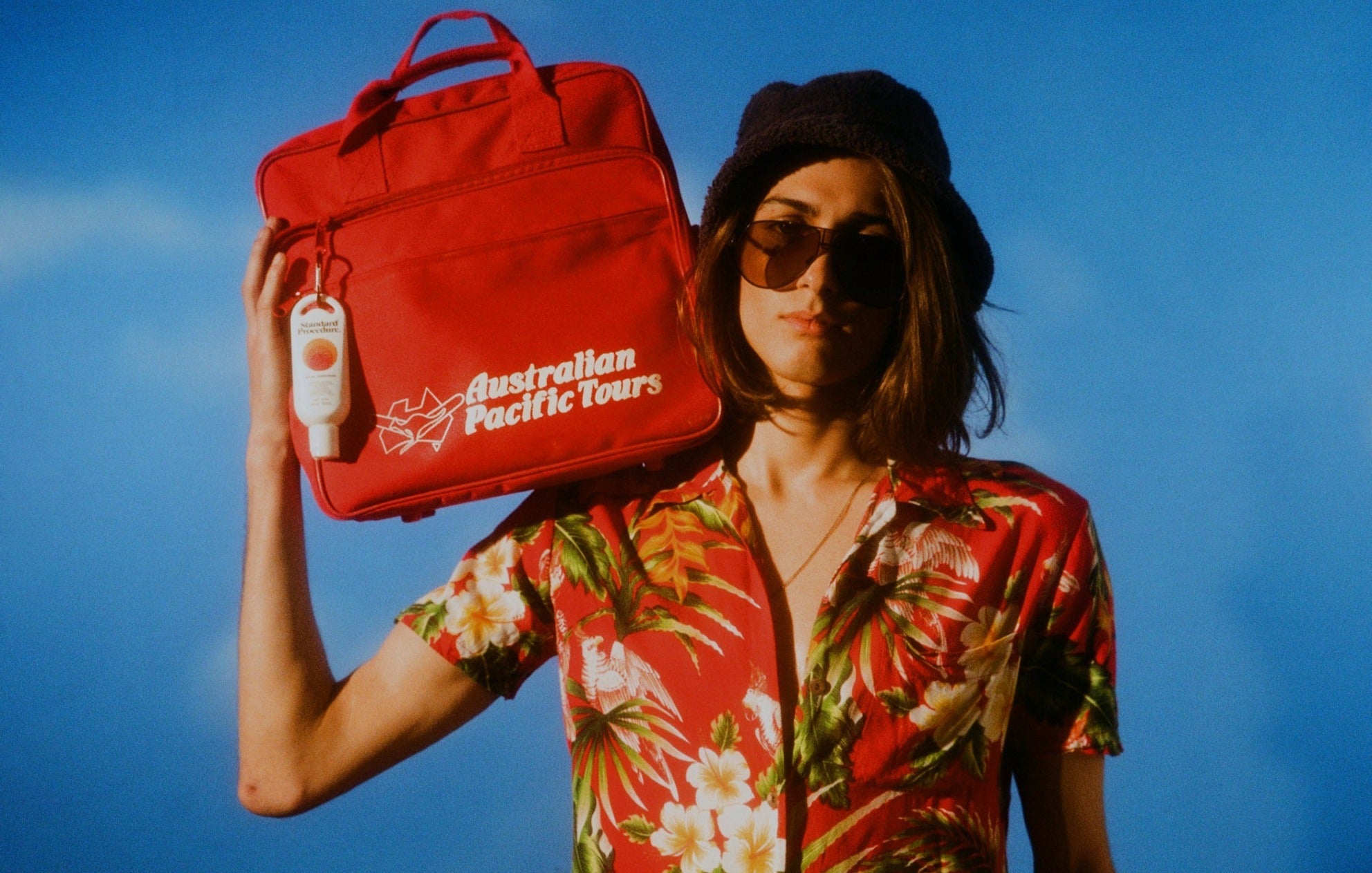 Person holding a red Australian Pacific Tours bag with a Standard Procedure Clip-On SPF 50+ clipped to it.
