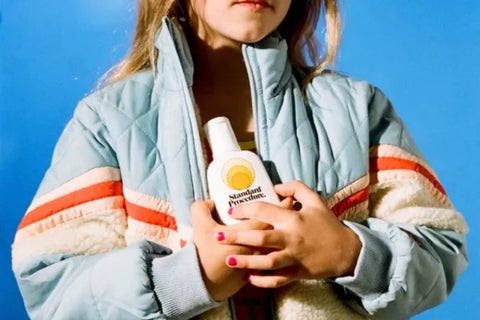 Antioxidant sunscreen bottle of Standard Procedure held by a child in a puffy jacket against a blue background.