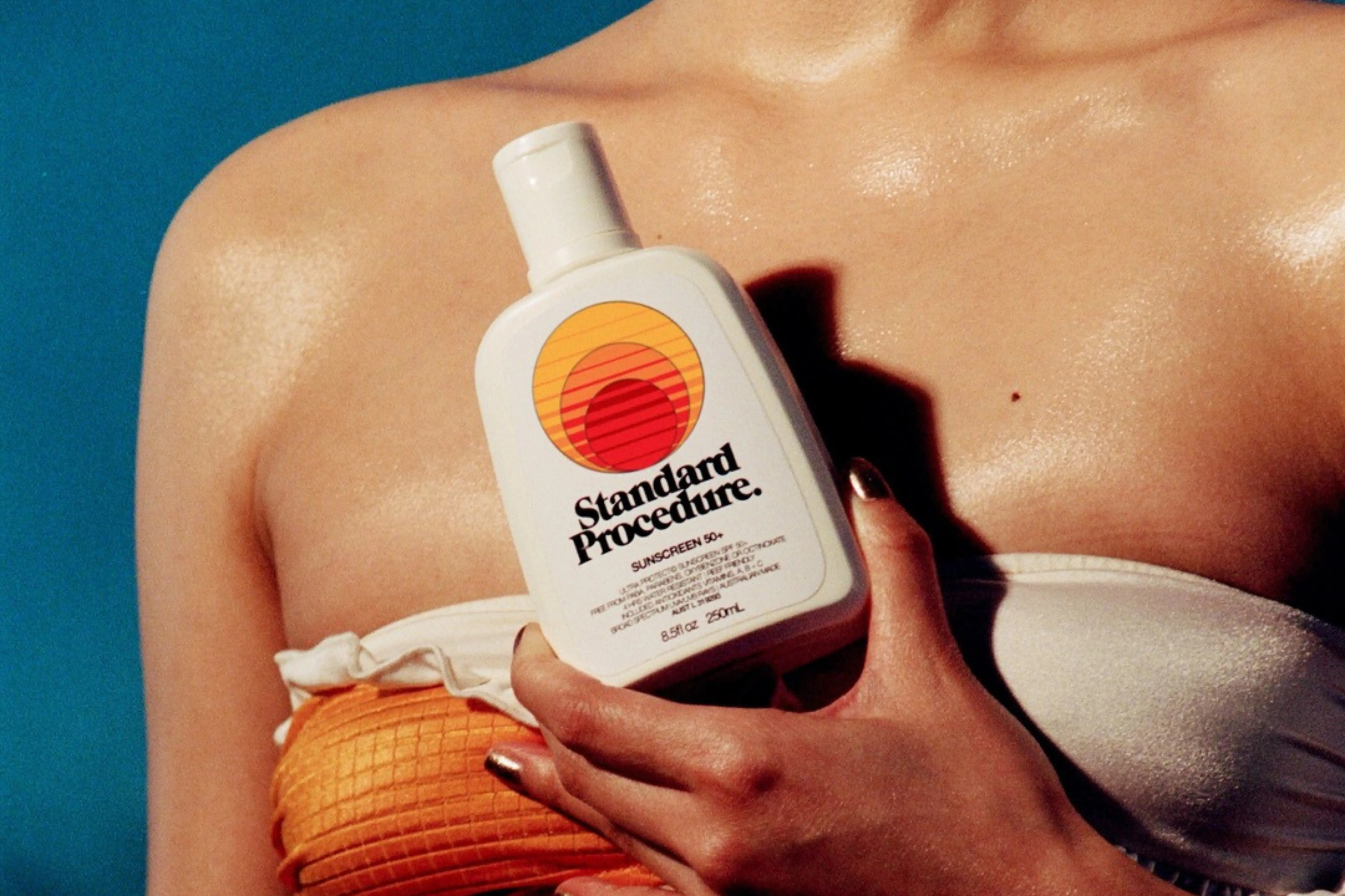 woman in bikini holding Standard Procedure daily sunscreen SPF 50+ bottle against blue background