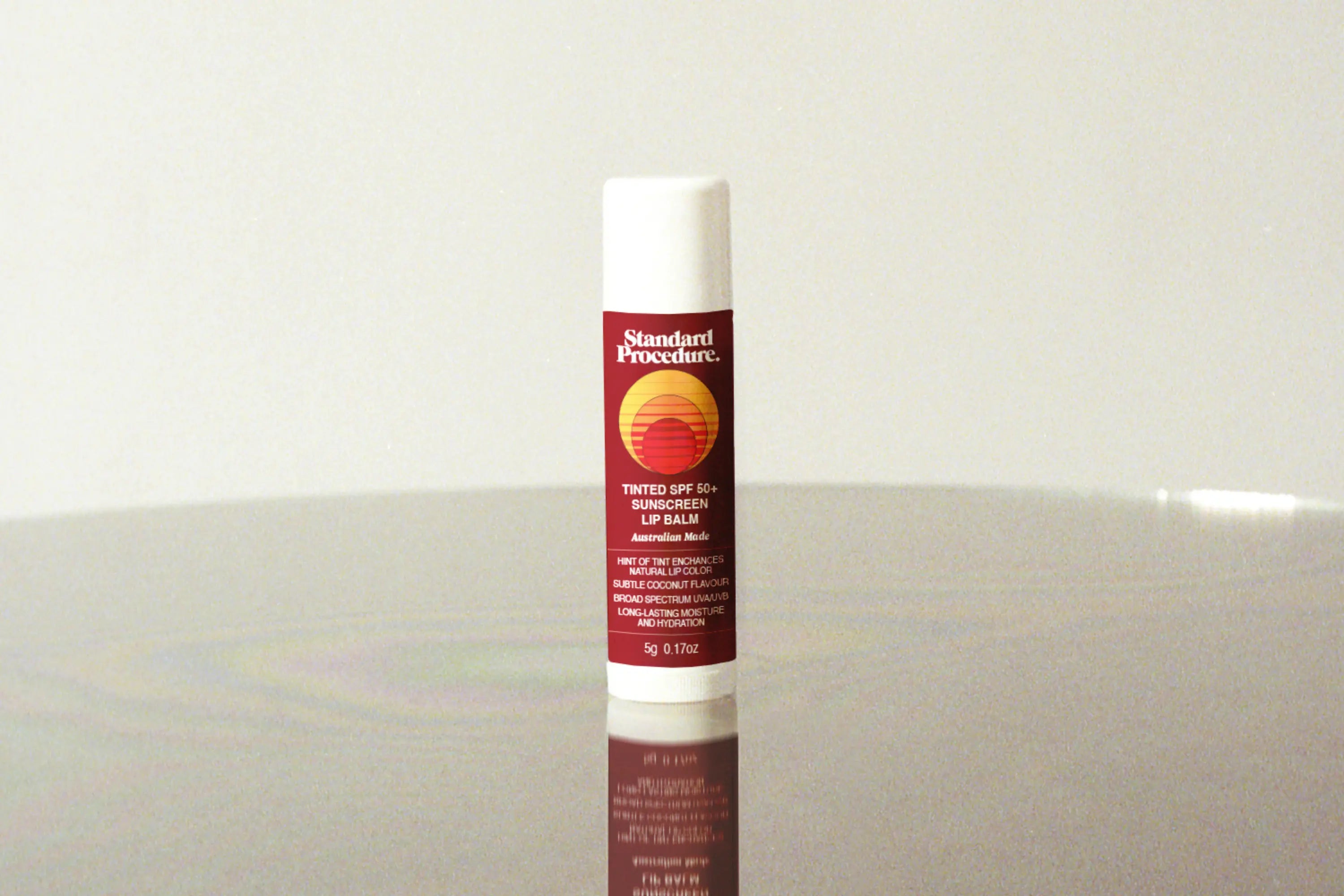 Lip balm with sunscreen SPF 50+ standing upright on a reflective surface against a neutral background