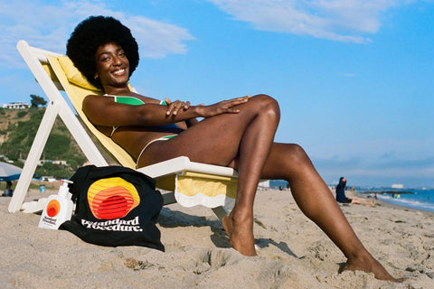 Non-greasy sunscreen beside a smiling woman sunbathing on a beach chair with a branded tote