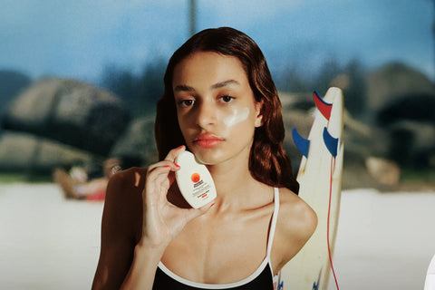 Sunscreen for face held by a woman at the beach with white cream on her cheek and a surfboard behind her.