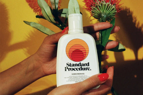 Water-resistant sunscreen bottle of Standard Procedure held in front of tropical flowers against a yellow background.