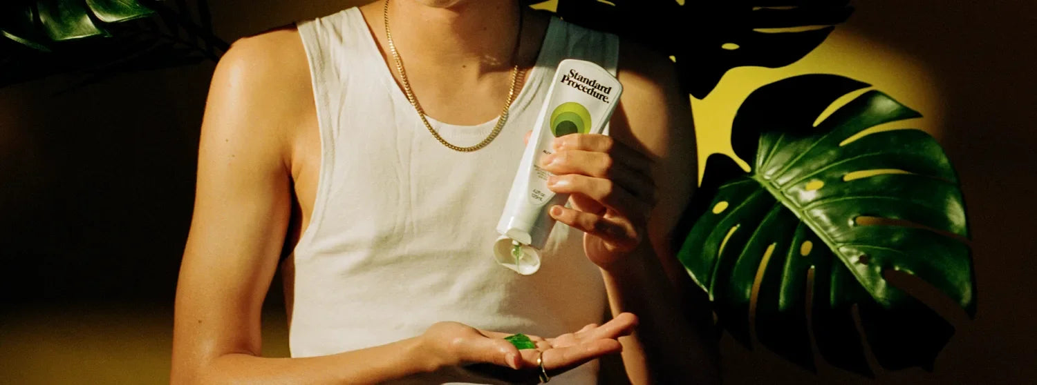 Person squeezing green Standard Procedure aloe vera gel into palm beside monstera leaves.