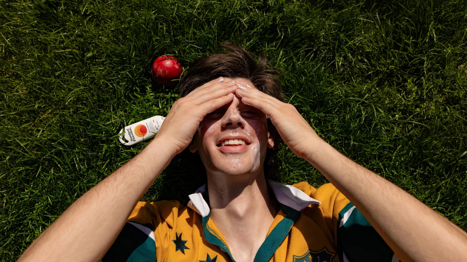Person rubbing Standard Procedure SPF 50+ sunscreen onto face while lying on grass beside a cricket ball.