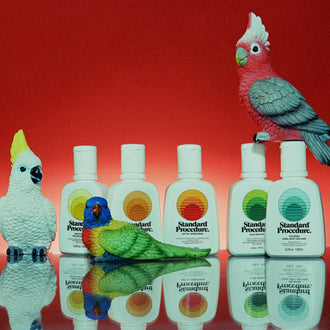 Australian mineral sunscreen bottles from Standard Procedure displayed alongside colorful Australian bird figurines against a red backdrop, highlighting vibrant, nature-inspired skincare