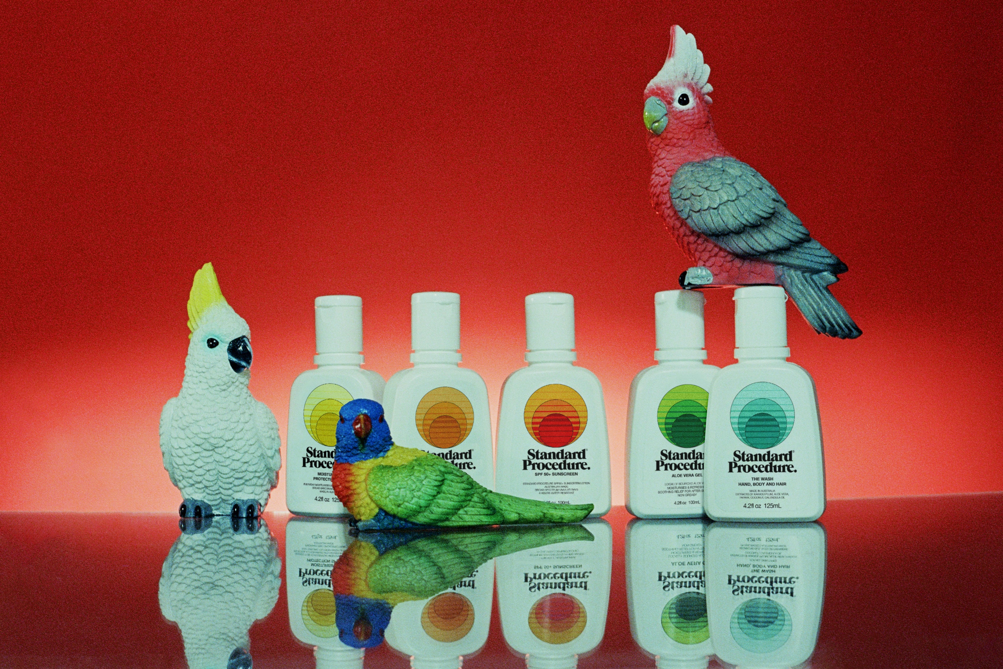 Australian mineral sunscreen bottles from Standard Procedure displayed alongside colorful Australian bird figurines against a red backdrop, highlighting vibrant, nature-inspired skincare