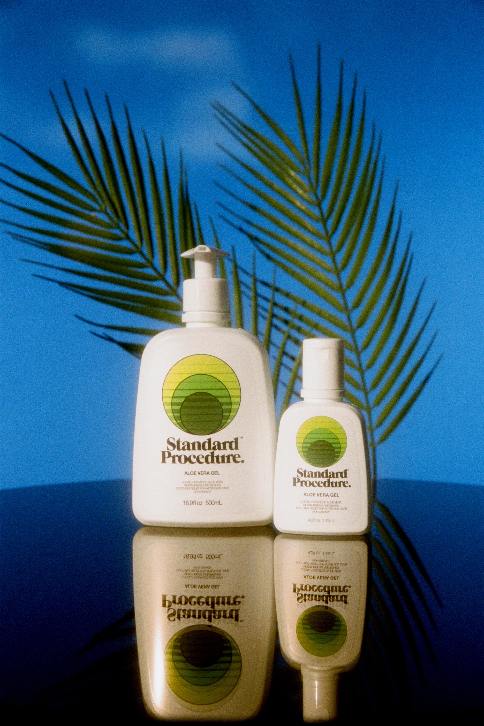 Aloe vera sun gel bottles from Standard Procedure displayed with tropical palm shadows and a reflective surface, emphasizing soothing, cooling after-sun skincare made with natural aloe