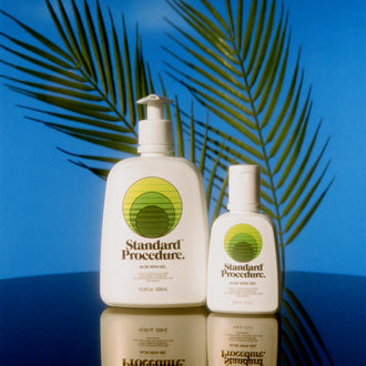 Aloe vera sun gel bottles from Standard Procedure displayed with tropical palm shadows and a reflective surface, emphasizing soothing, cooling after-sun skincare made with natural aloe