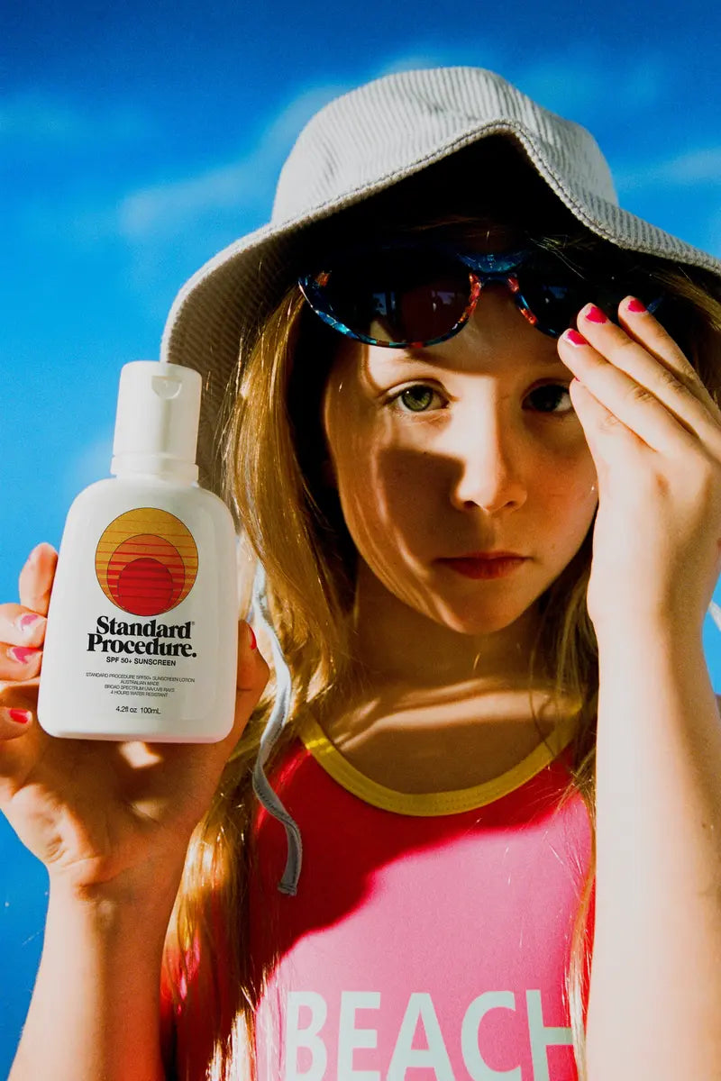 Person in bucket hat holding Standard Procedure SPF 50+ travel sunscreen bottle at the beach.