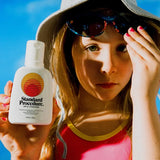 Person in bucket hat holding Standard Procedure SPF 50+ travel sunscreen bottle at the beach.