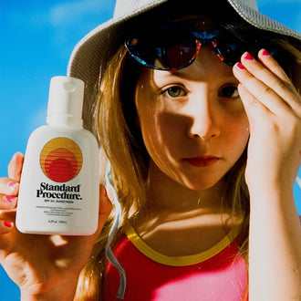 Person in bucket hat holding Standard Procedure SPF 50+ travel sunscreen bottle at the beach.
