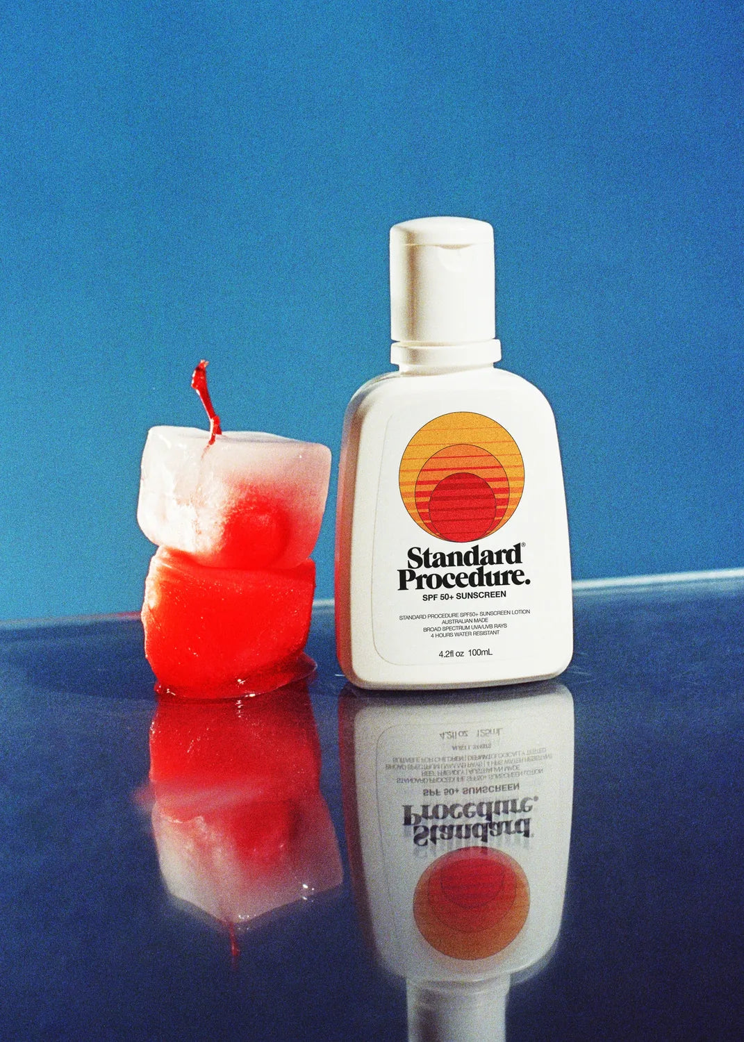 Standard Procedure SPF 50+ sunscreen 100ml travel bottle beside melting ice on a blue reflective surface.