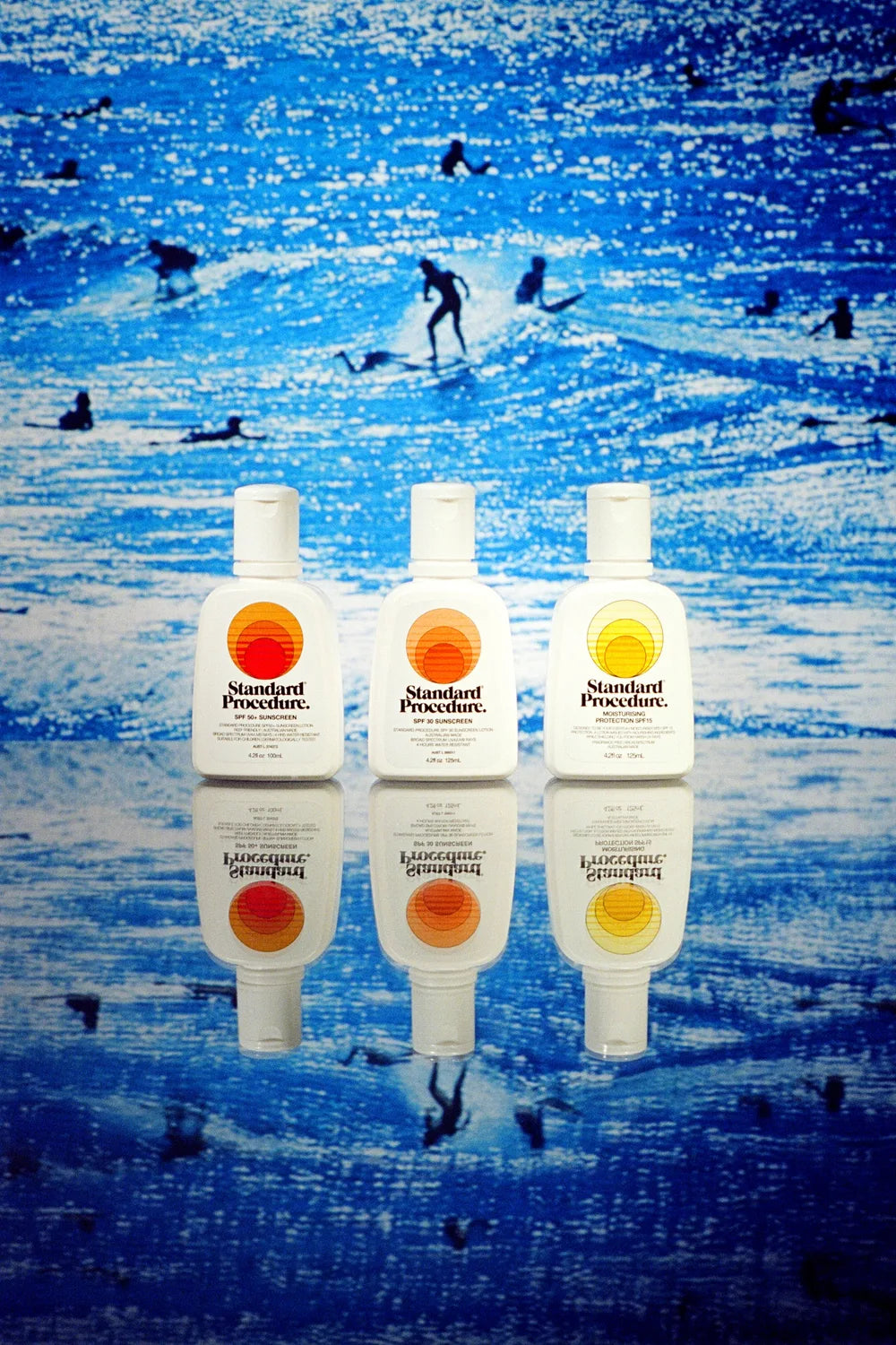 Standard Procedure SPF 50+, SPF 30, and SPF 15 sunscreens lined up against a surf scene.