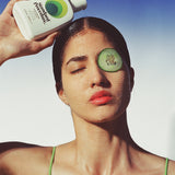 Woman holding a Standard Procedure Aloe Vera 250ml bottle with cucumber slices on her face against a blue sky.