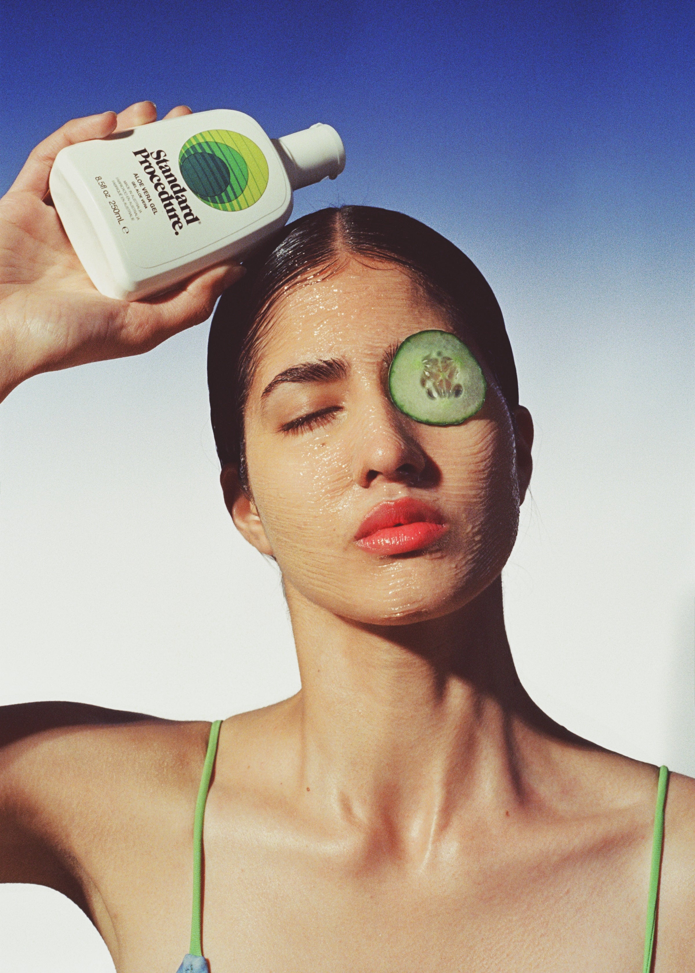Woman holding a Standard Procedure Aloe Vera 250ml bottle with cucumber slices on her face against a blue sky.