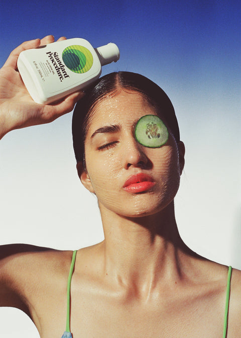 Woman holding a Standard Procedure Aloe Vera 250ml bottle with cucumber slices on her face against a blue sky.