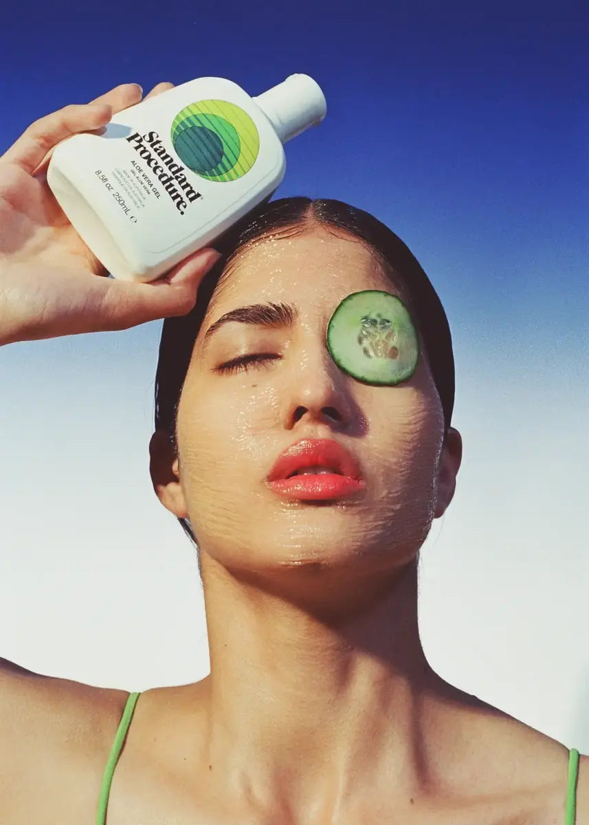 Woman holding a Standard Procedure Aloe Vera 250ml bottle with cucumber slices on her face against a blue sky.