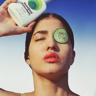 Woman holding a Standard Procedure Aloe Vera 250ml bottle with cucumber slices on her face against a blue sky.