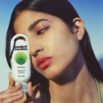 Close-up of a woman holding a Standard Procedure Aloe Vera Gel 60ml tube against a clear blue sky.