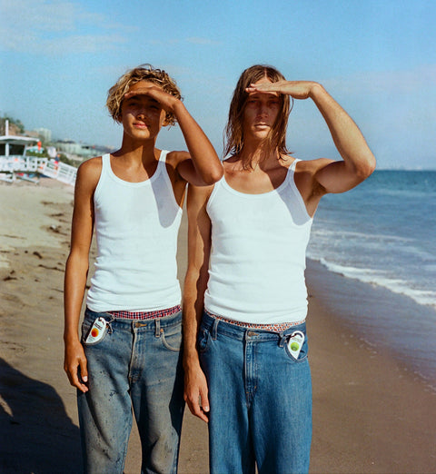 Two people wearing white tank tops and blue jeans on a beach with Sunscreen clipped into their belt buckle