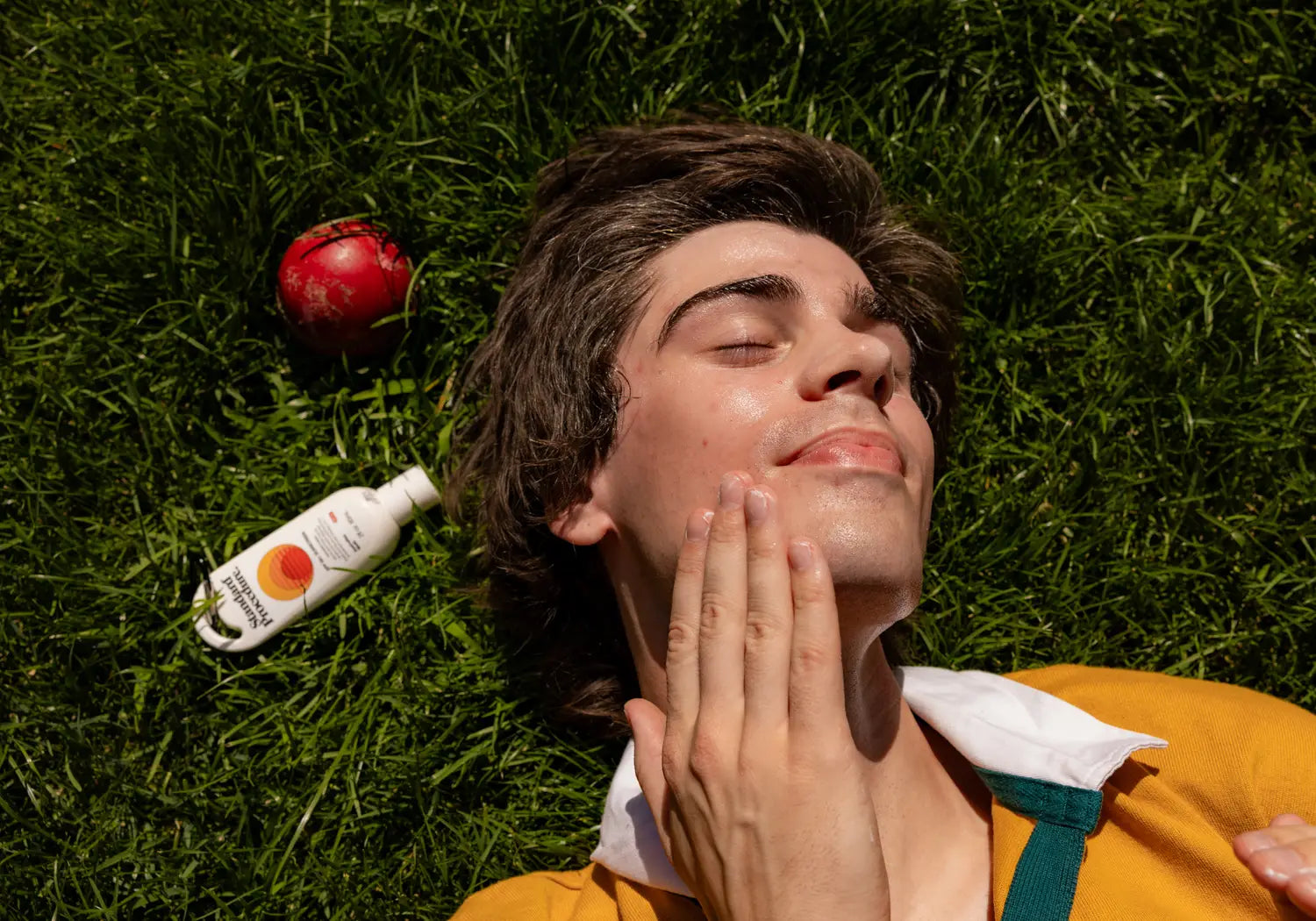 Person lying on grass applying Standard Procedure SPF 50 sunscreen to their face, cricket ball nearby.