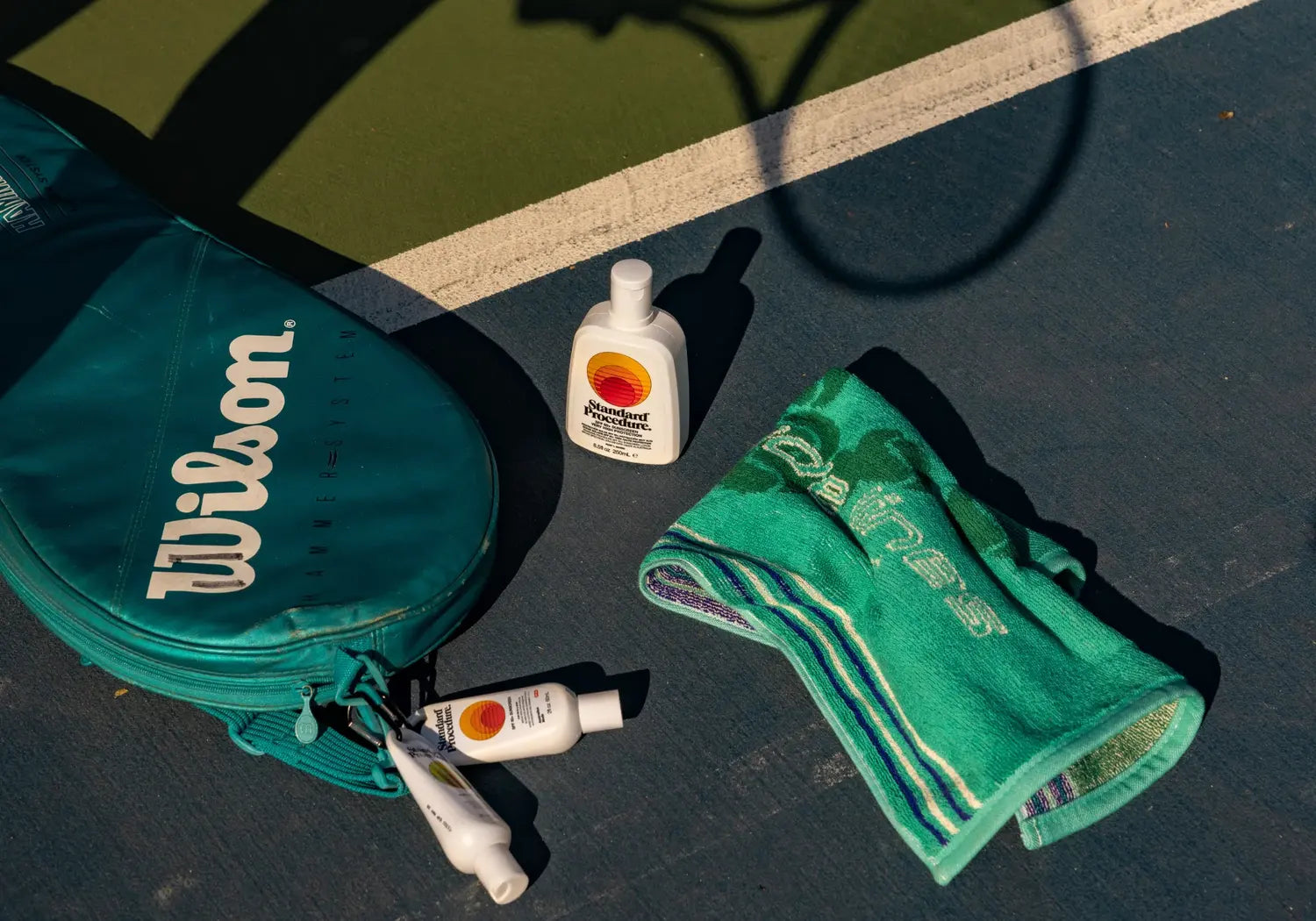 Standard Procedure SPF 50 sunscreen bottles beside a Wilson tennis bag and green court towel.