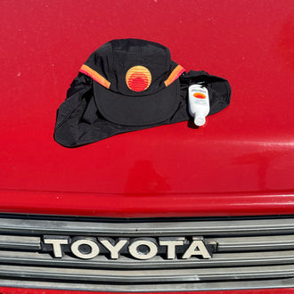 Black surf cap featuring the Standard Procedure logo placed on a red surface beside a small bottle of sunscreen, emphasizing stylish and practical sun protection essentials