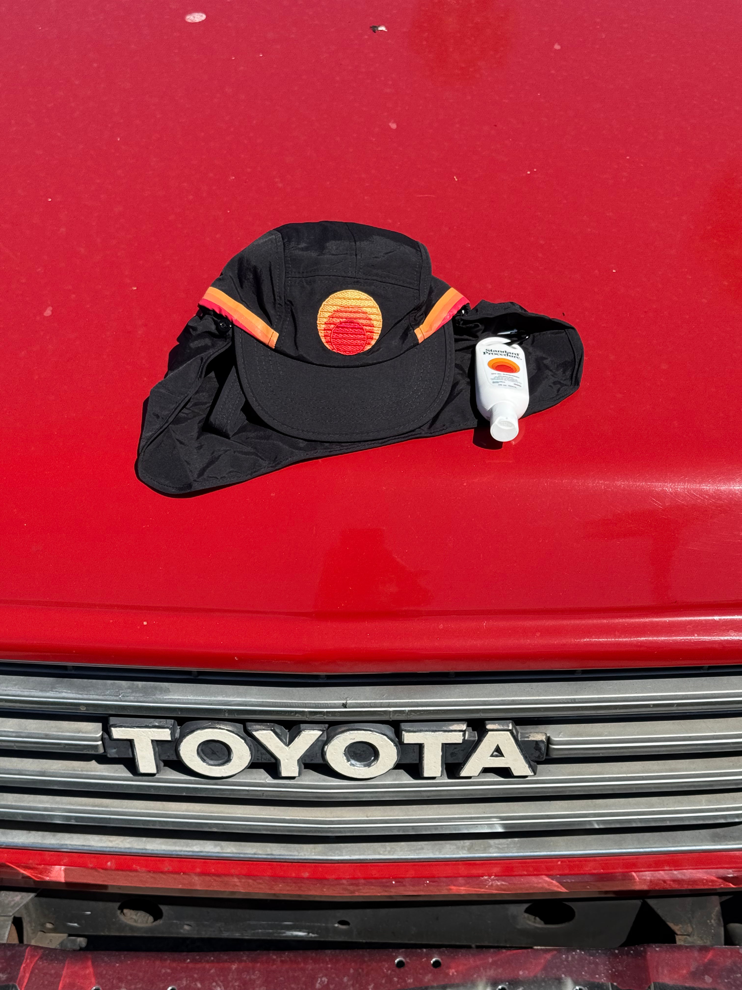 Black surf cap featuring the Standard Procedure logo placed on a red surface beside a small bottle of sunscreen, emphasizing stylish and practical sun protection essentials