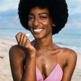 Smiling beachgoer in a pink bikini holding a Standard Procedure SPF 50 lip balm by the shore.