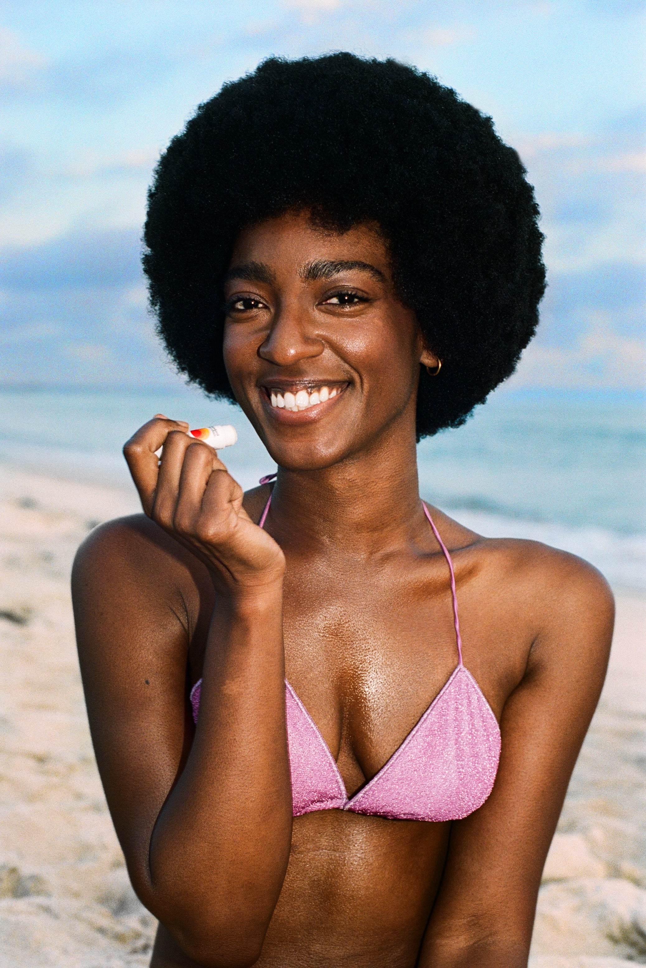 Smiling beachgoer in a pink bikini holding a Standard Procedure SPF 50 lip balm by the shore.