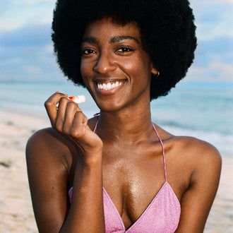 Smiling beachgoer in a pink bikini holding a Standard Procedure SPF 50 lip balm by the shore.