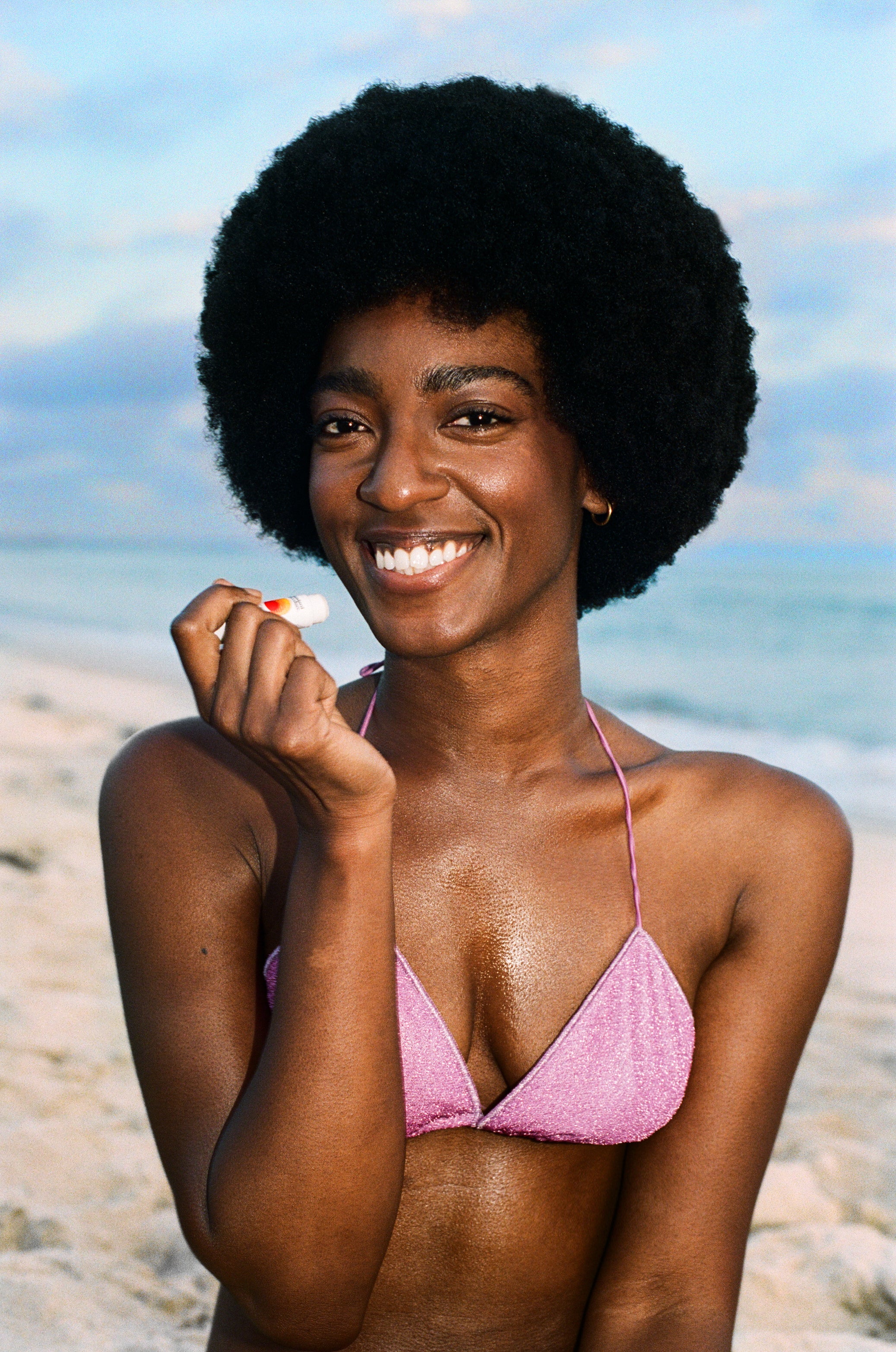 Smiling beachgoer in pink bikini holding Standard Procedure SPF 50 lip balm by the ocean.