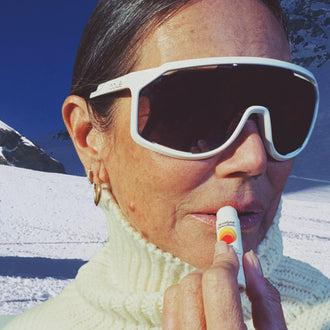 Woman applying Standard Procedure SPF 50 Lip Balm on a snowy mountain wearing white ski goggles and a turtleneck.