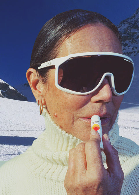 Woman applying Standard Procedure SPF 50 Lip Balm on a snowy mountain wearing white ski goggles and a turtleneck.