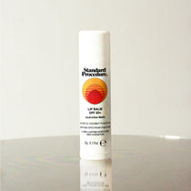 Standard Procedure Lip Balm SPF 50 in coconut flavour with the sunset logo on a reflective surface.