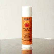 Standard Procedure orange zinc SPF 50+ sunscreen stick with retro sunset label on reflective surface.