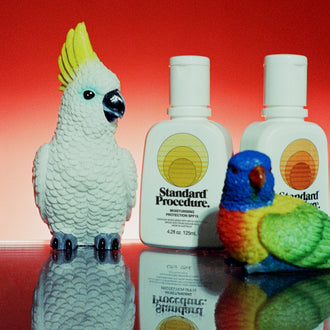 Two Standard Procedure SPF 15 moisturiser bottles styled with cockatoo and lorikeet figurines on red.
