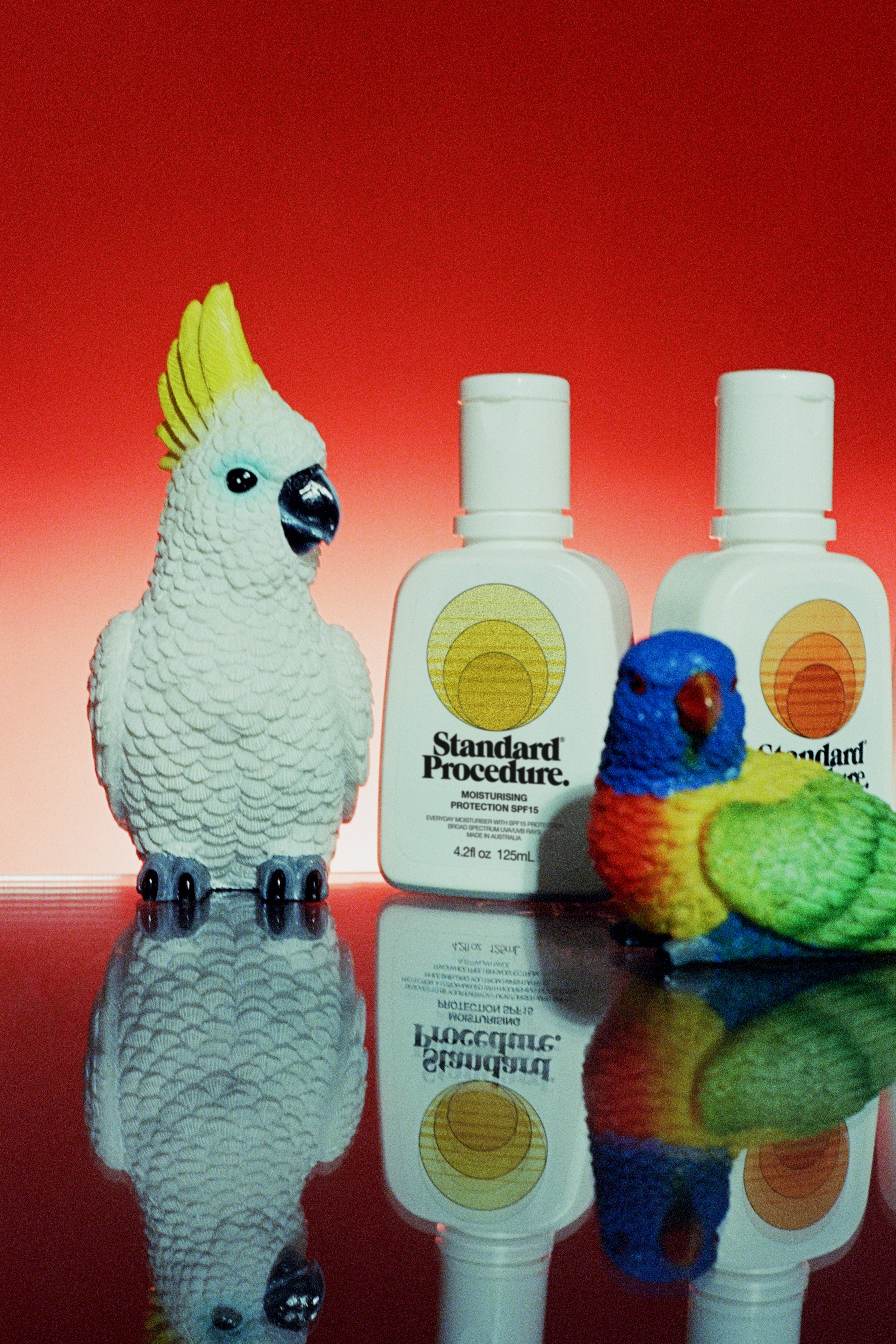 Two Standard Procedure SPF 15 moisturiser bottles styled with cockatoo and lorikeet figurines on red.